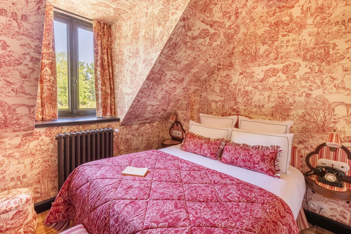Une chambre avec un plafond en pente, un lit recouvert d’une courtepointe à motifs floraux rouges, une fenêtre avec des rideaux et un téléphone ancien posé sur une table de chevet.