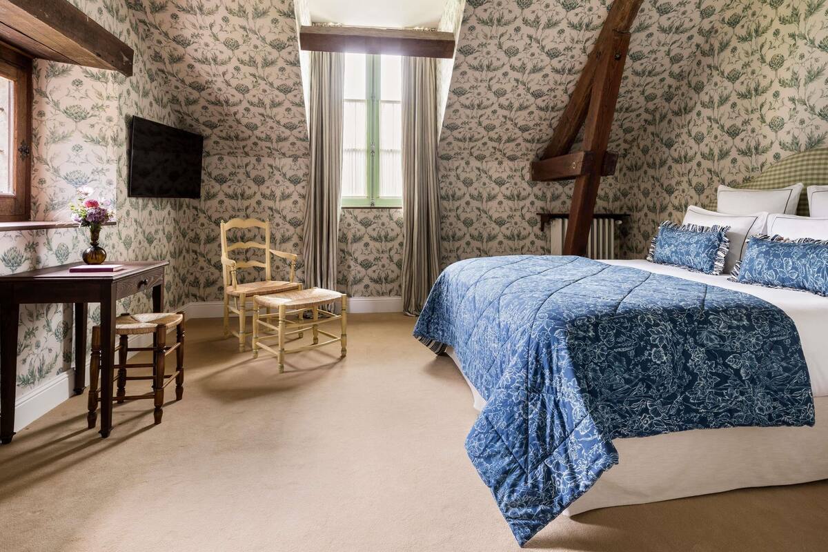 A bedroom with floral wallpaper, a bed with blue floral bedding, a wooden chair, a small table with a vase, and a window with curtains.