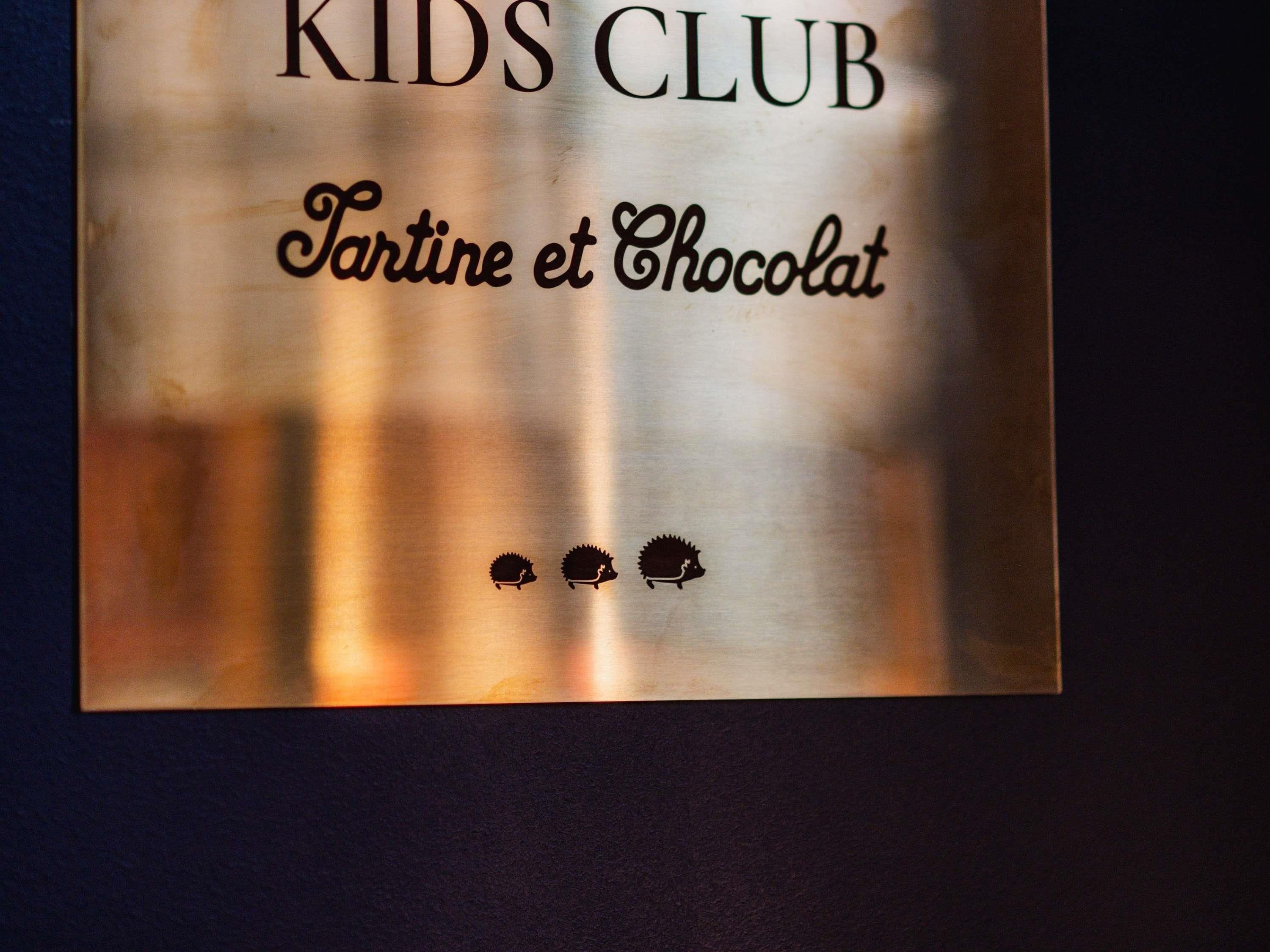 Un senyal metàl·lic amb el text "KIDS CLUB" i "Tartine et Chocolat", acompanyat de dos icones de eriçons.