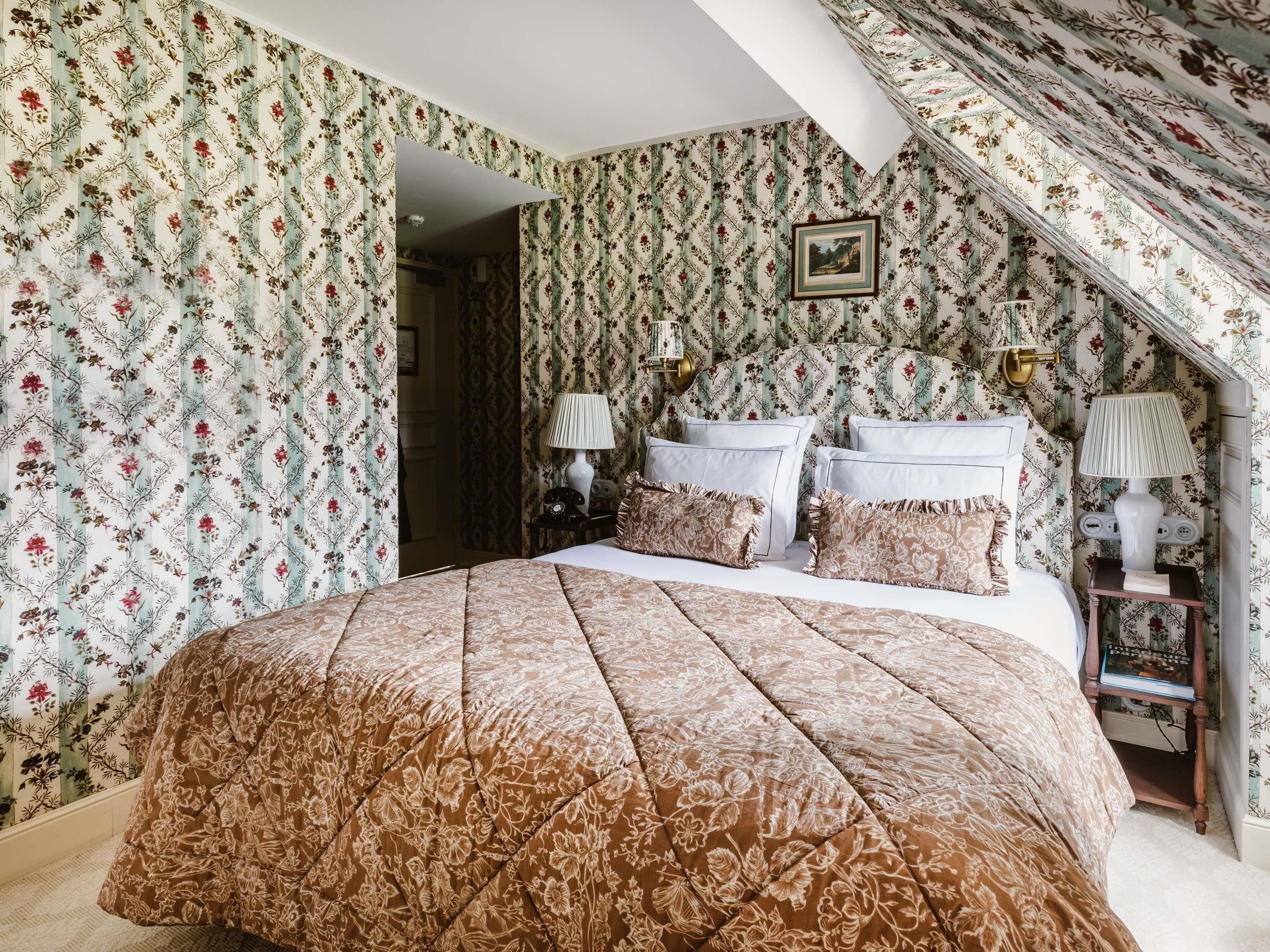 A bedroom with floral wallpaper, a large bed with a patterned duvet, two bedside tables with lamps, and a framed picture on the wall.