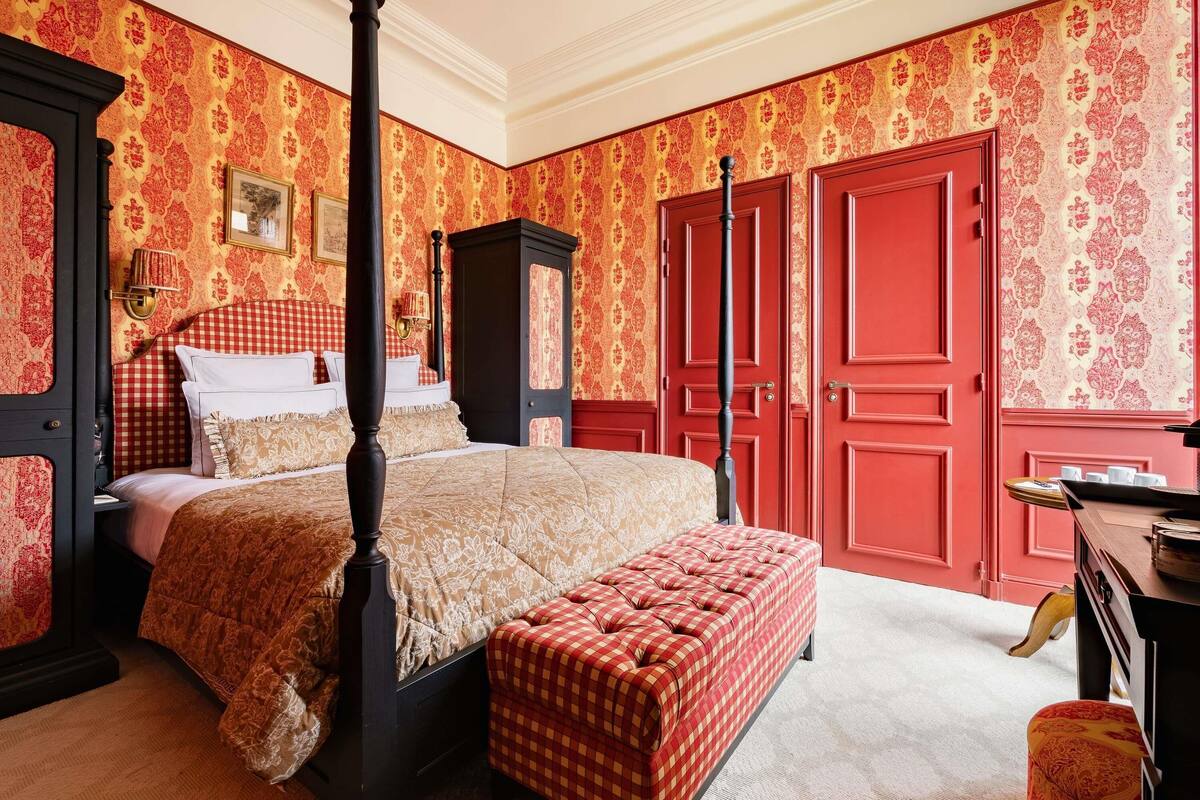 A four-poster bed with a checkered ottoman, red doors, and patterned wallpaper.