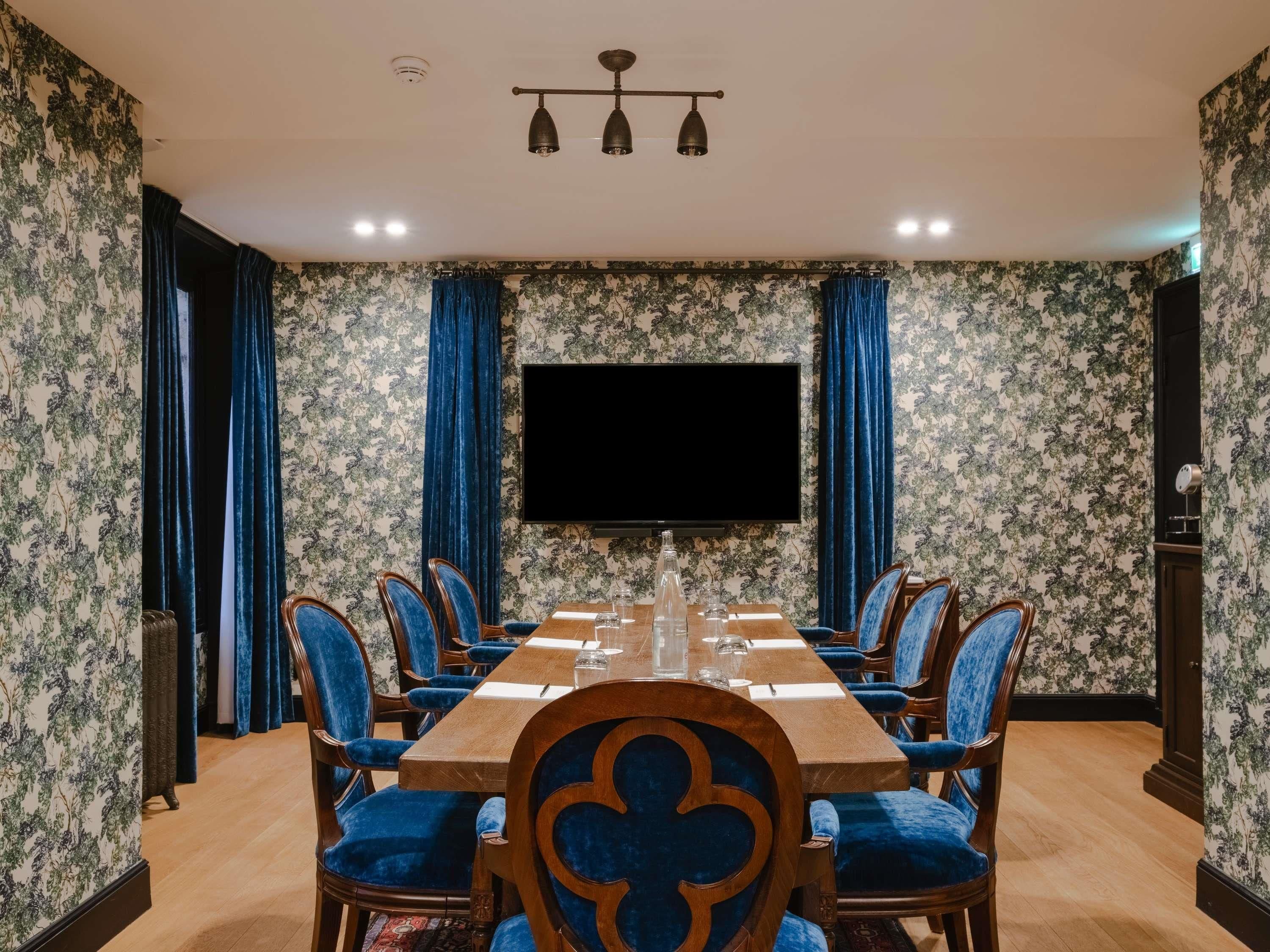 A conference room with a long table, blue upholstered chairs, a flat-screen TV, and floral wallpaper.