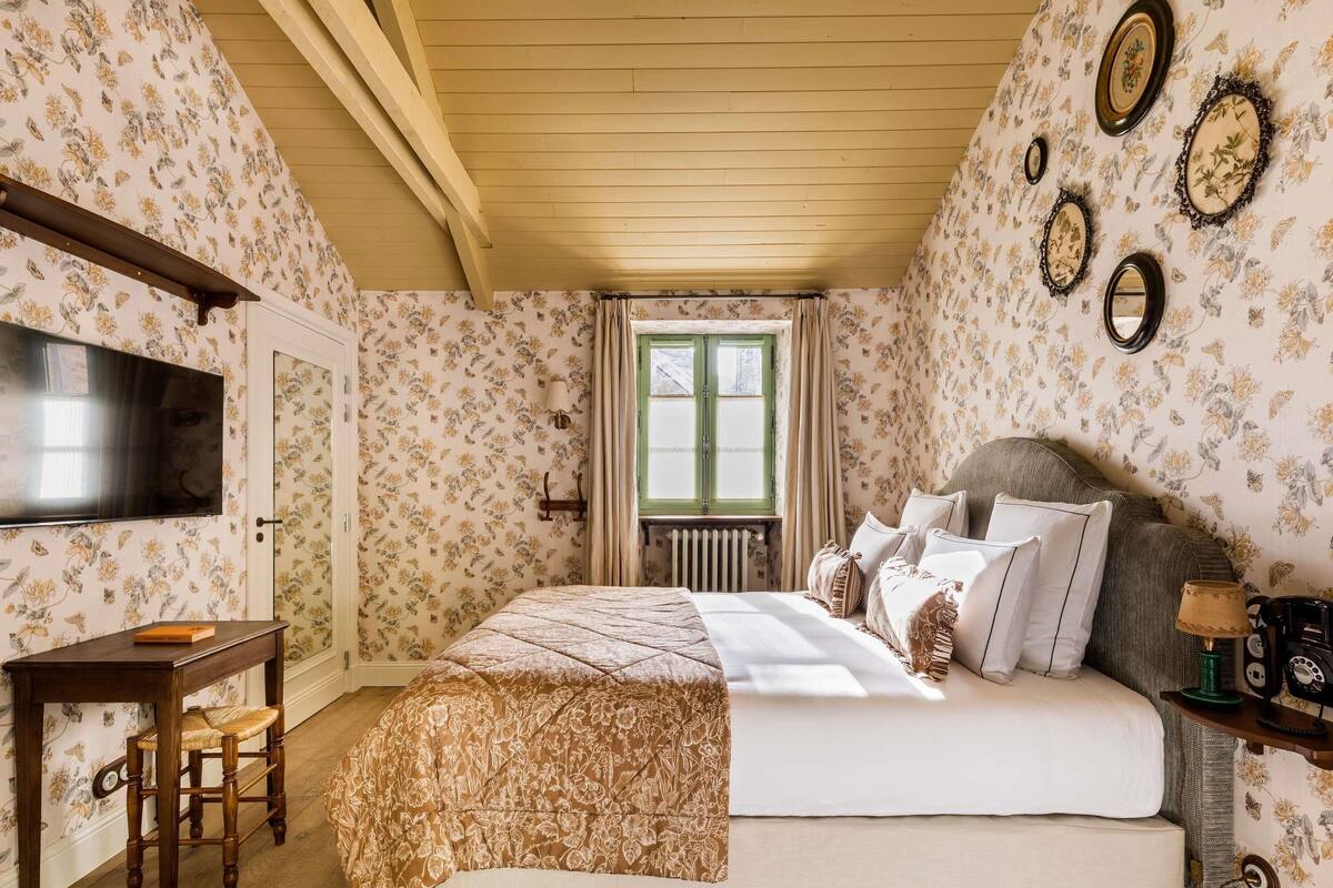 A bedroom with a bed, a desk, a chair, a TV, and a window with curtains.
