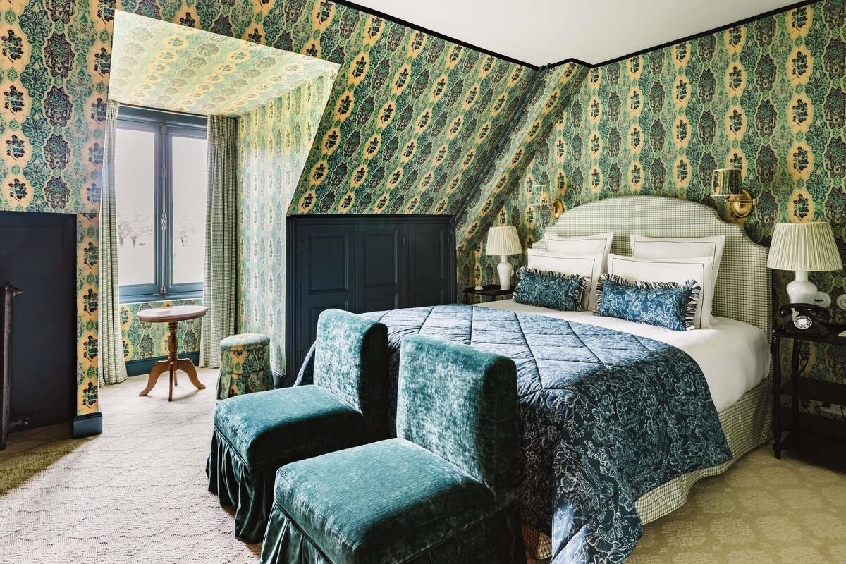 A bedroom with a large bed, two matching armchairs, a small round table, and a window with curtains.