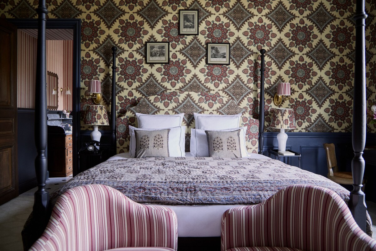 Een hemelbed met een beddengoed met patroon, twee nachtkastjes met lampen, en een spiegel boven de kaptafel.