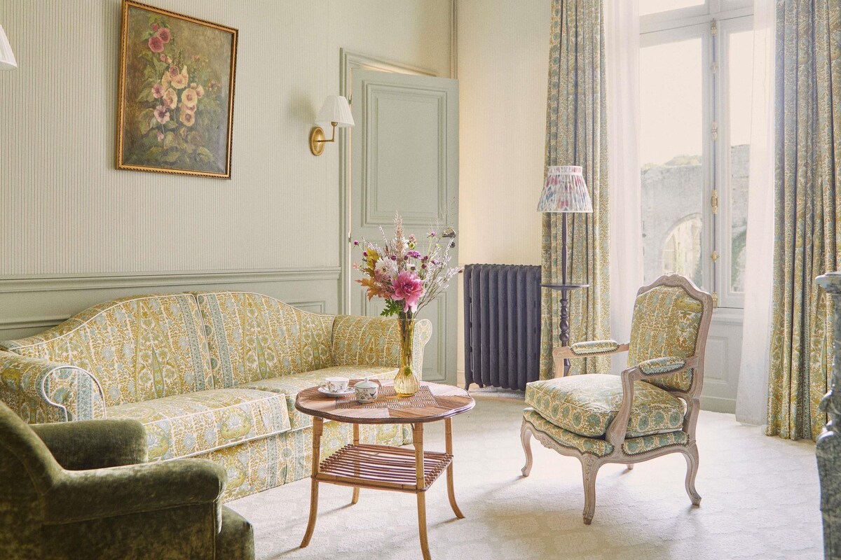 Una habitación con un sofá estampado, una silla, una mesita con un jarrón, una l ámpara y vistas al exterior por una ventana.