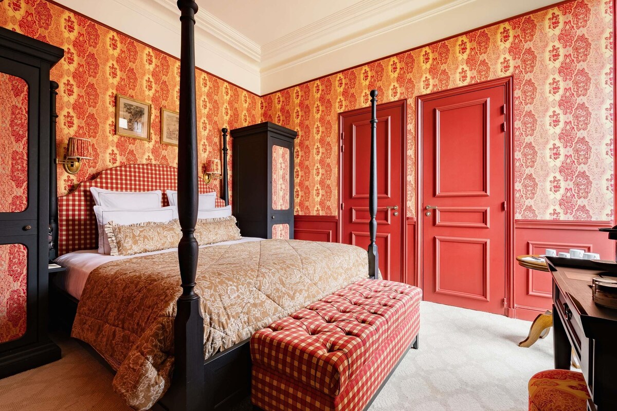 Un lit à baldaquin avec un pouf à carreaux, des portes rouges et un papier peint à motifs.