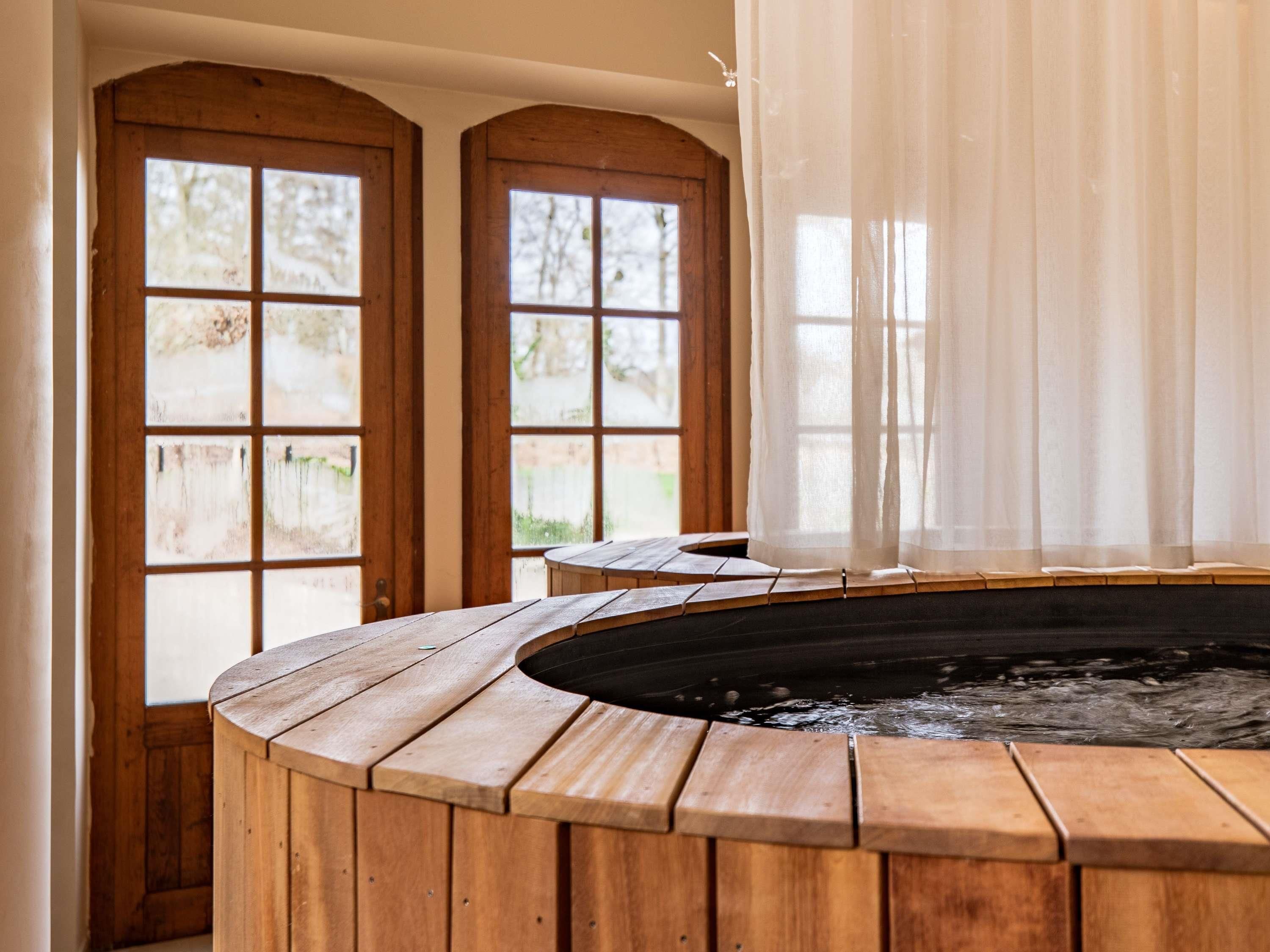 A wooden hot tub with a view of a garden through large windows.
