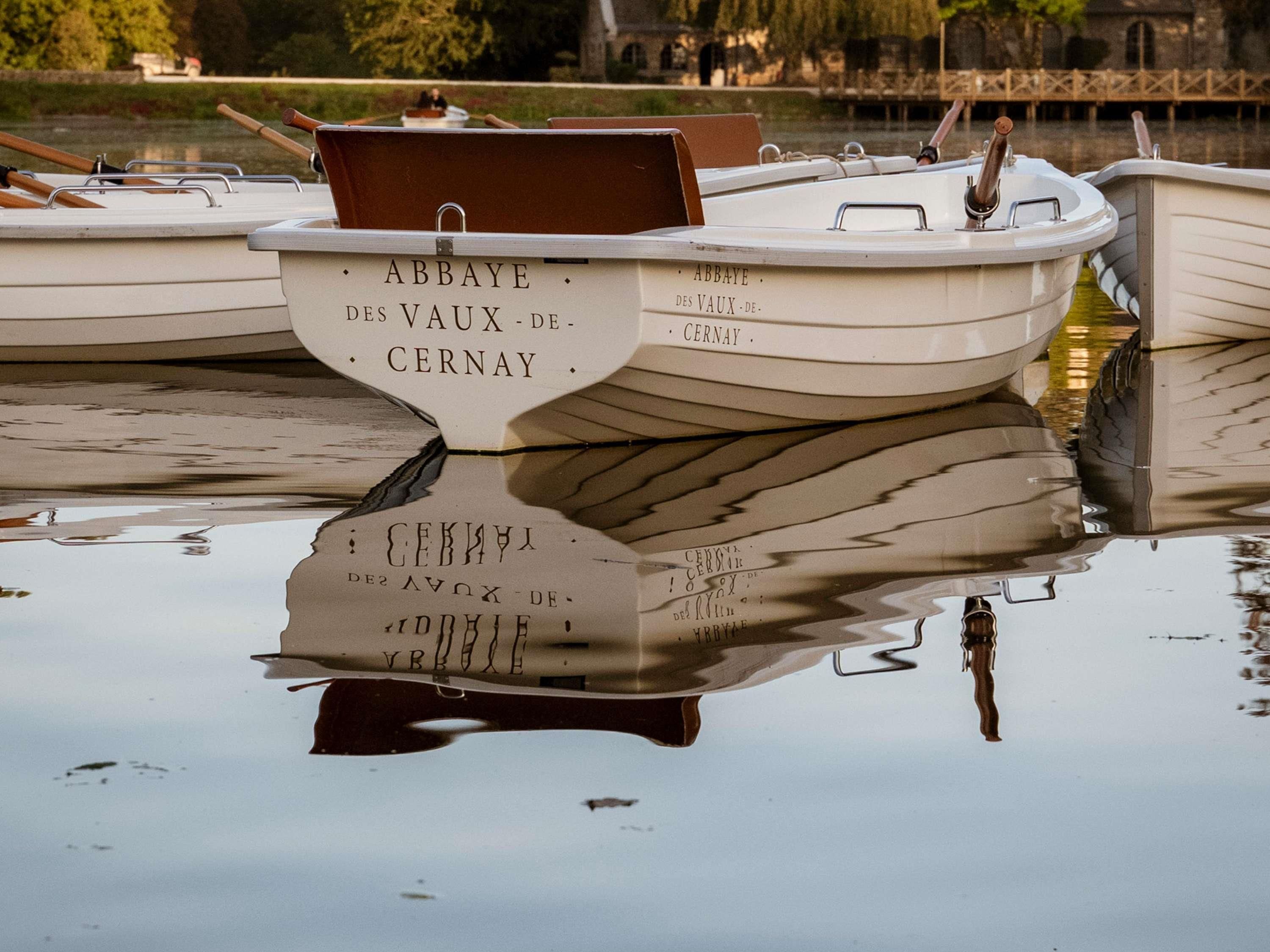 A row of boats docked at a calm body of water, with 'ABBAYE DES VAUX-DE-CERNAY' written on the side of one boat.
