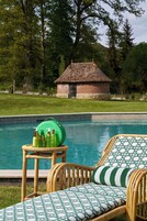 Uma área ao lado da piscina com móveis de vime e uma mesa com garrafas, um edifício de tijolos ao fundo e um gramado.