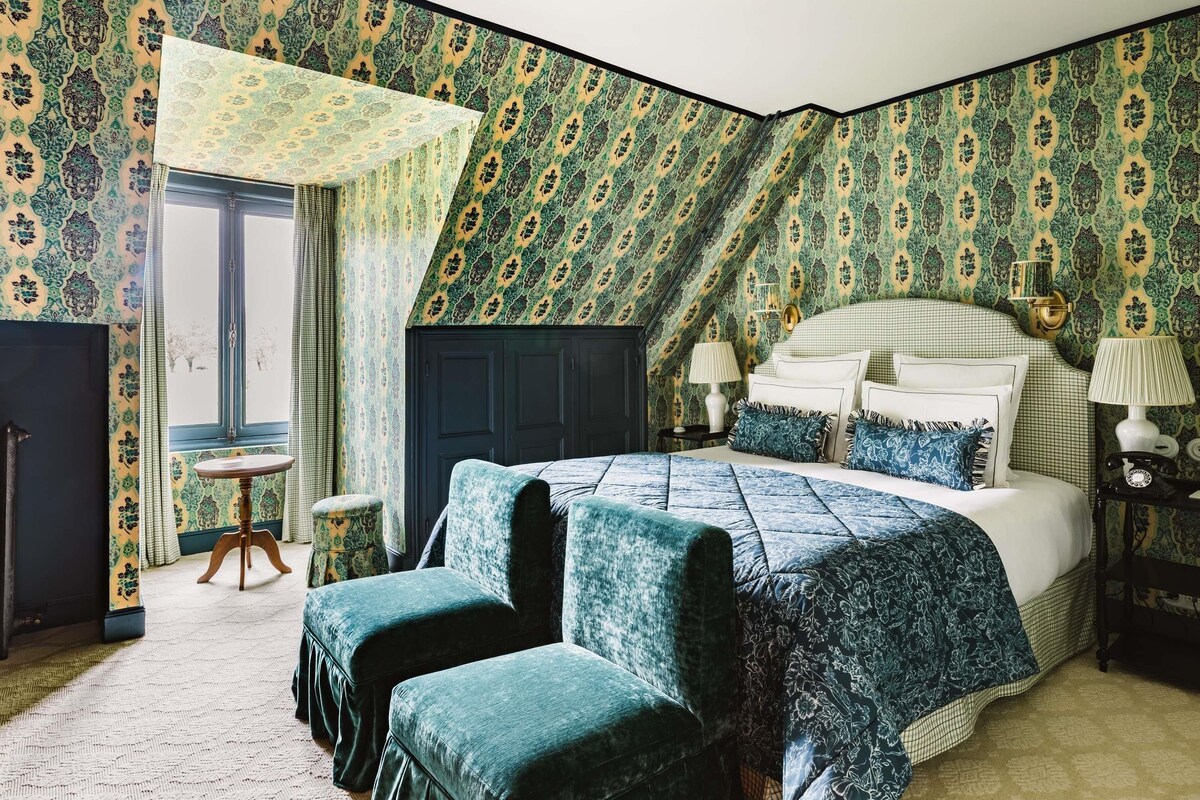 Une chambre à coucher avec un grand lit, deux fauteuils assortis, une petite table ronde et une fenêtre avec des rideaux.