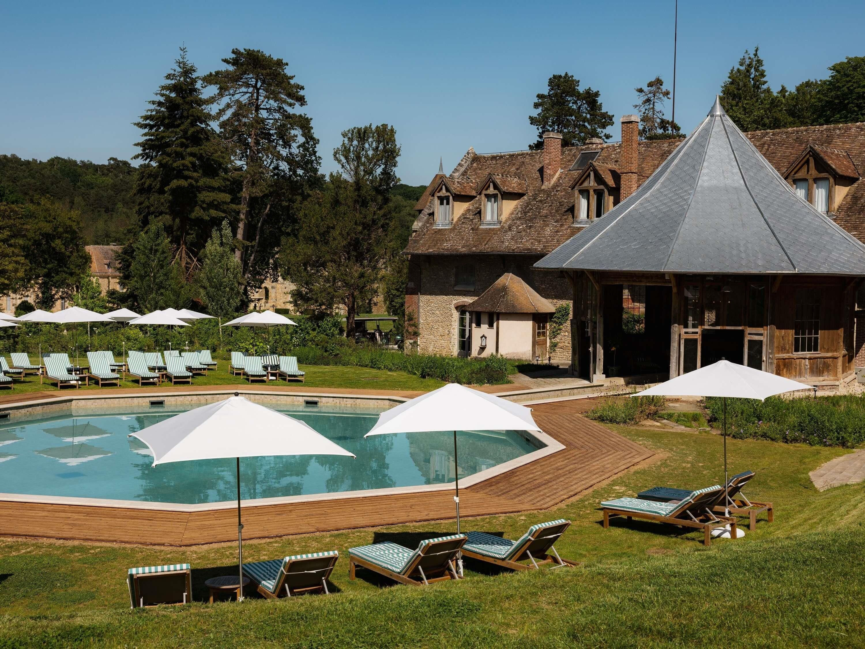 A swimming pool with lounge chairs and umbrellas, surrounded by a wooden deck and a building with a steep roof.