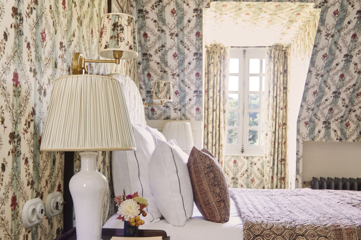 Een slaapkamer met bloemenbehang, een witte lamp, een nachtkastje met een boek, en een raam met gordijnen.