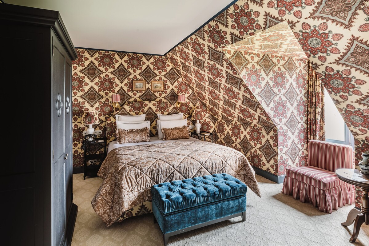 Een slaapkamer met een groot bed, een blauwe poef, een stoel en behang met een patroon.