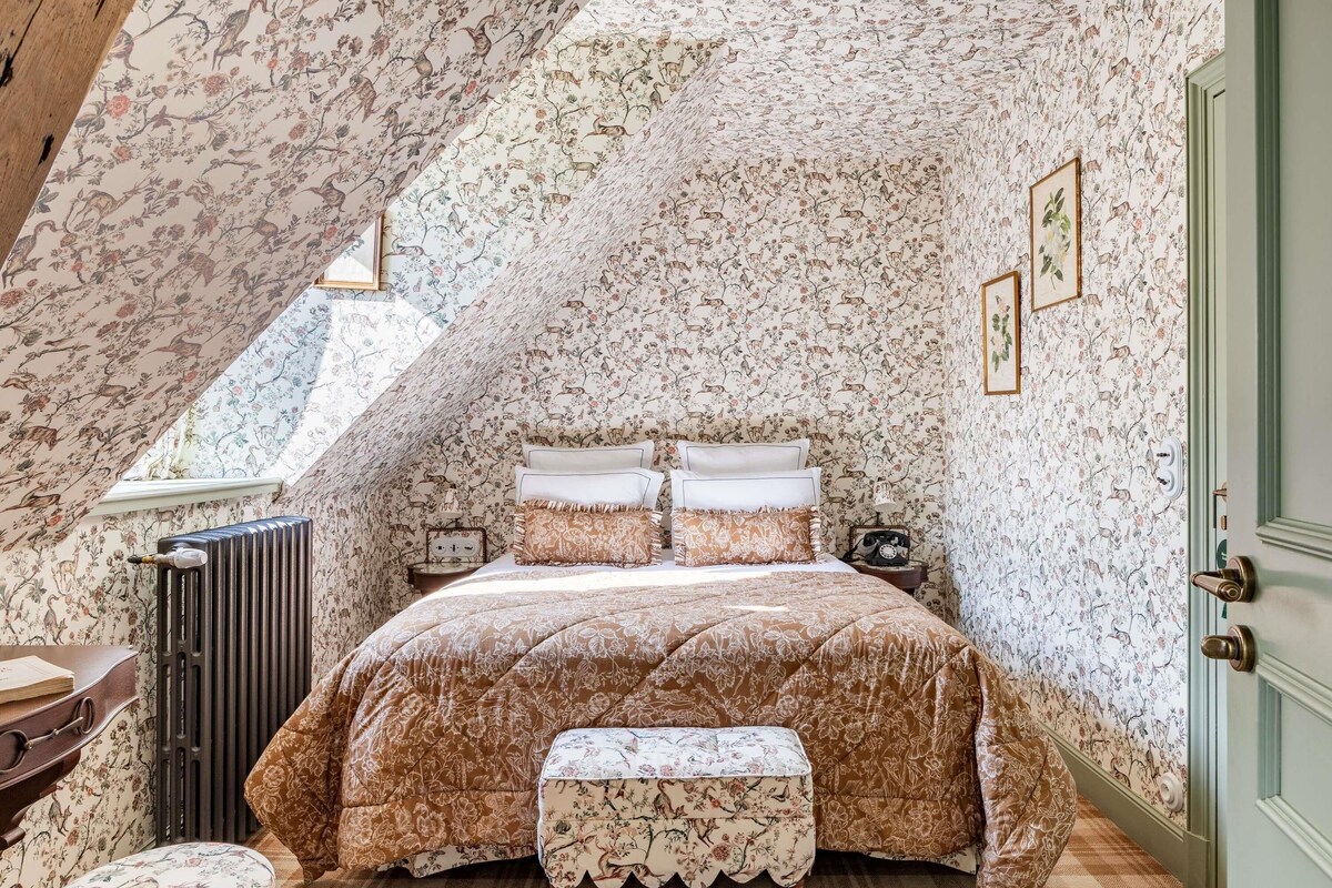 Een slaapkamer met een schuine plafond, bloemenbehang, een bed met bijpassende kussens, een radiator, een kleine tafel en een bankje.
