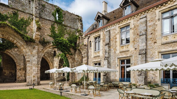 Une cour extérieure agrémentée d’arcades en pierre, un bâtiment doté de nombreuses fenêtres et des espaces de détente en plein air avec des parasols.