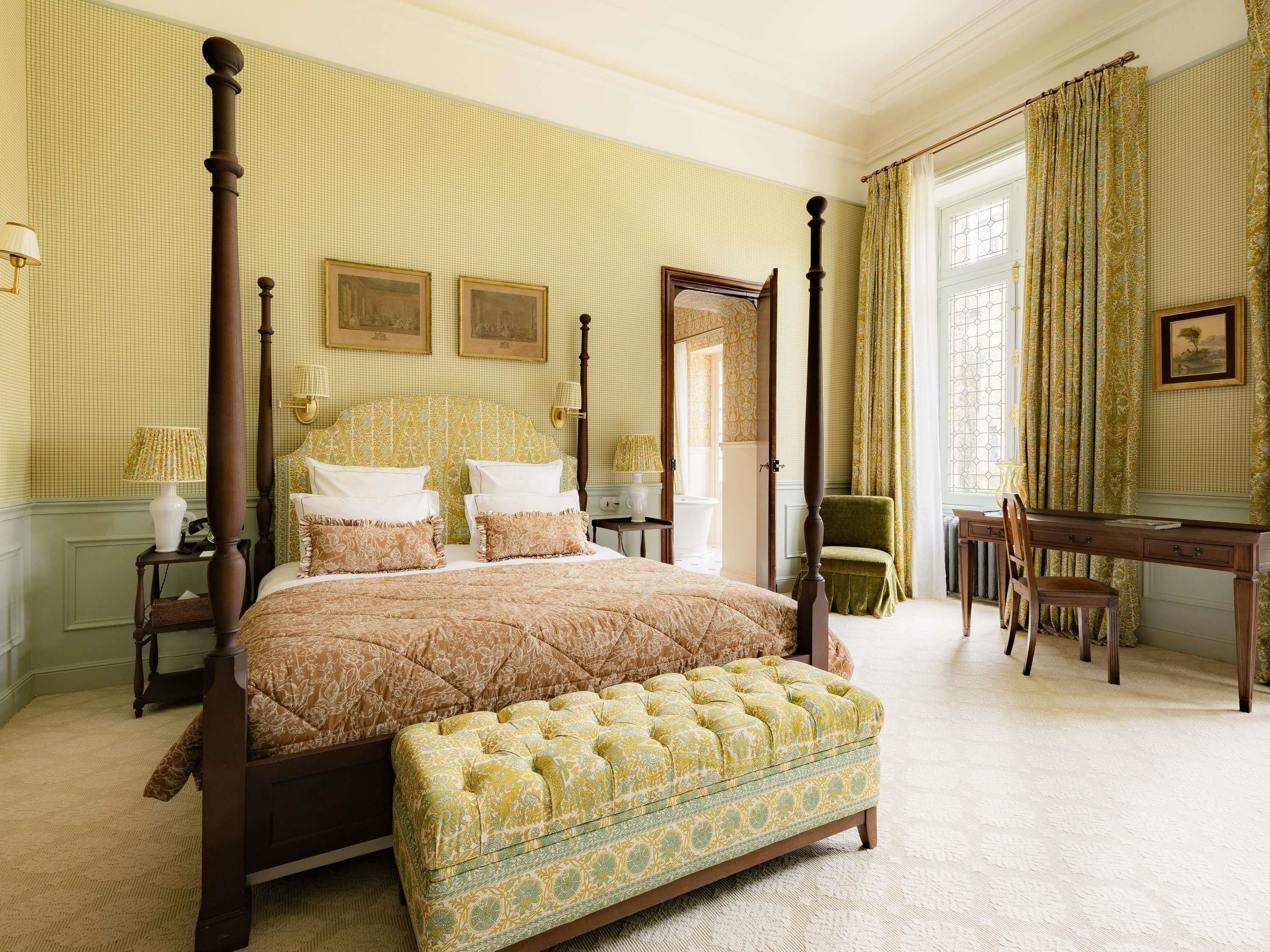 A four-poster bed with a tufted ottoman, a desk with a chair, and a view of a window with curtains.