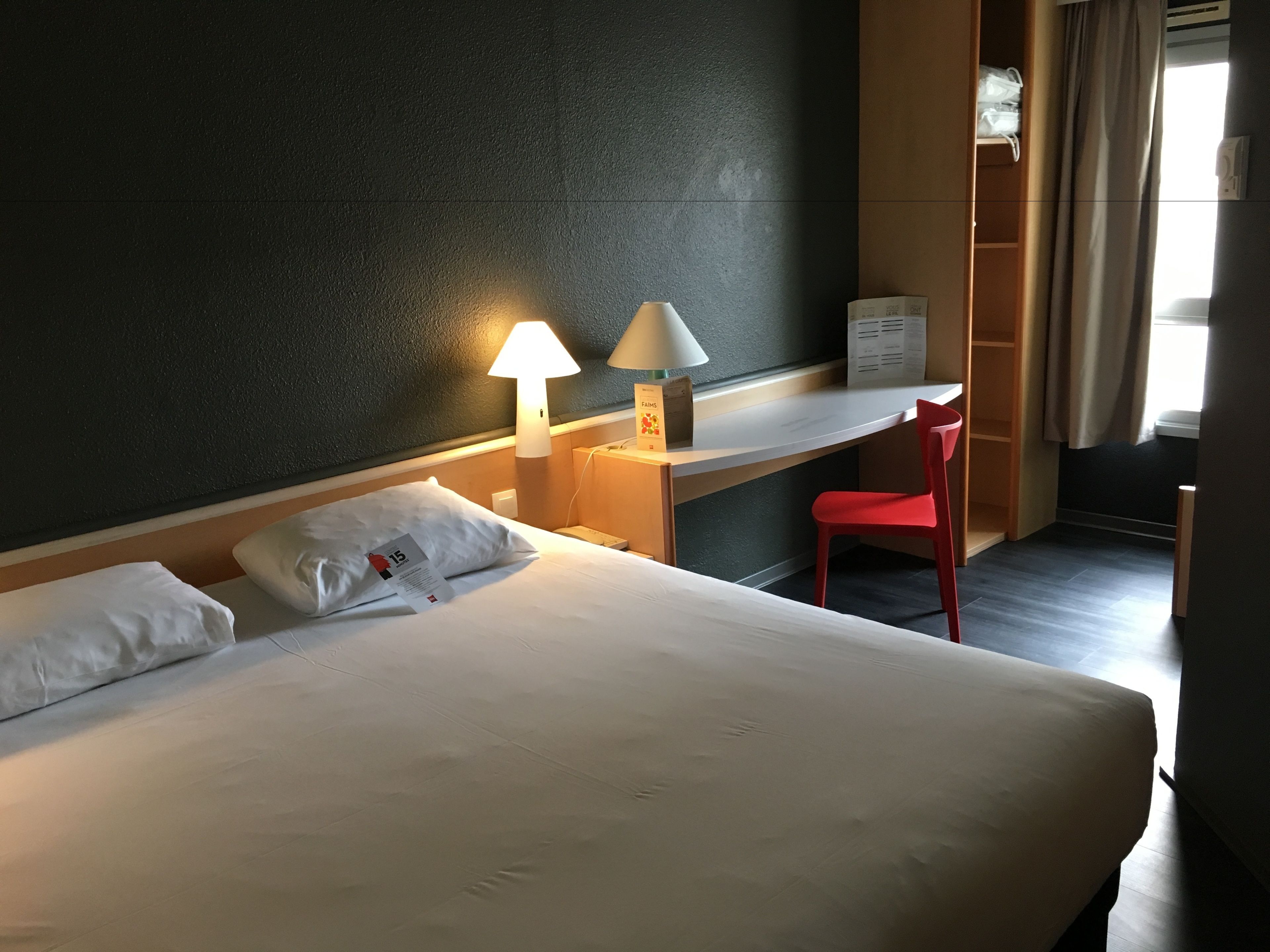 A hotel room with a bed, two bedside lamps, a desk, a red chair, and a window with curtains.