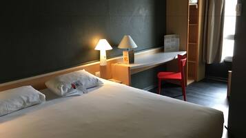 A hotel room with a bed, two bedside lamps, a desk, a red chair, and a window with curtains.