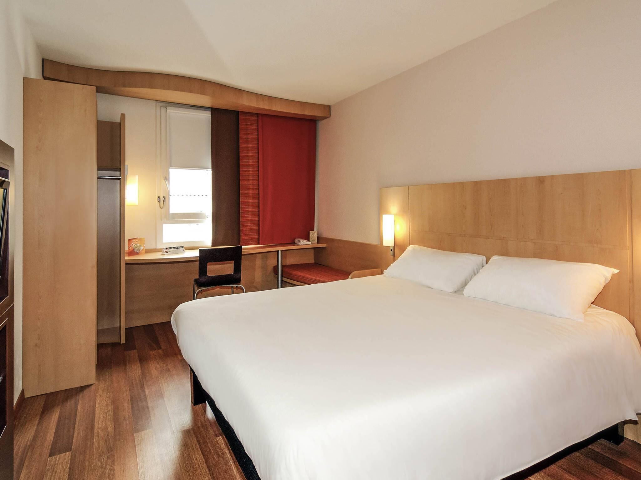A hotel room with a large bed, a desk with a chair, a mirror, and a window with curtains.