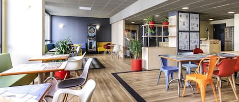 Un espacio interior moderno con varias zonas de estar, que incluyen mesas, sillas, un dardo y plantas en macetas.