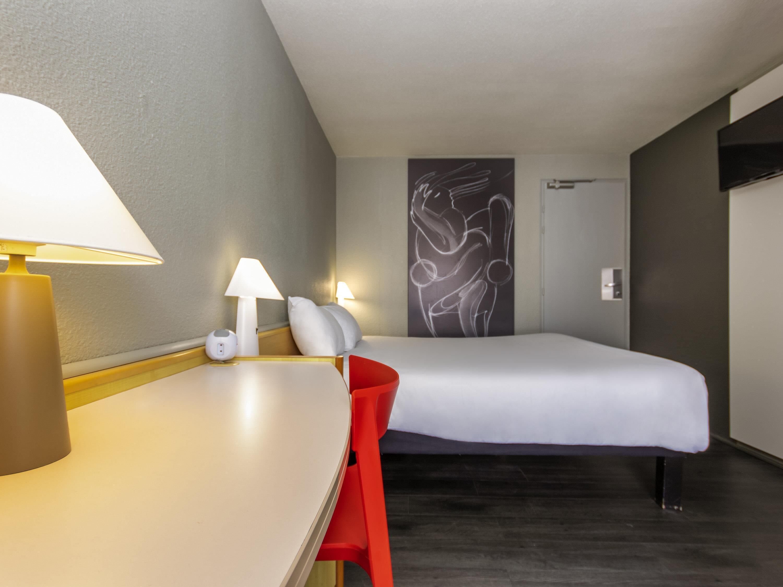 A hotel room with a bed, bedside lamps, a desk, a chair, and a wall art piece.