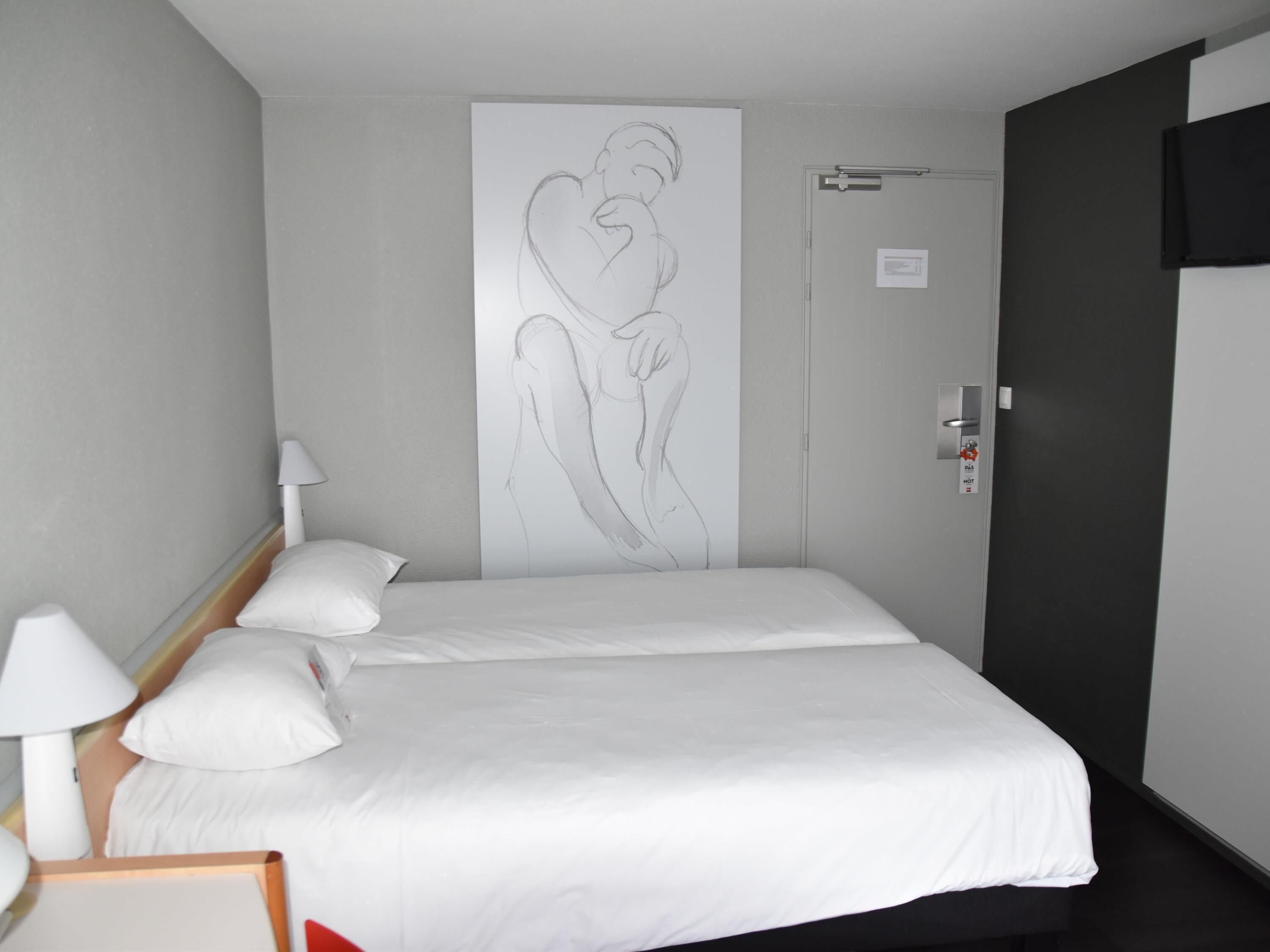 Ein Hotelzimmer mit zwei Betten, einer weißen Wand mit Herzzeichnung, einer Tür mit Rauchmelder und einem an der Wand befestigten Fernseher.