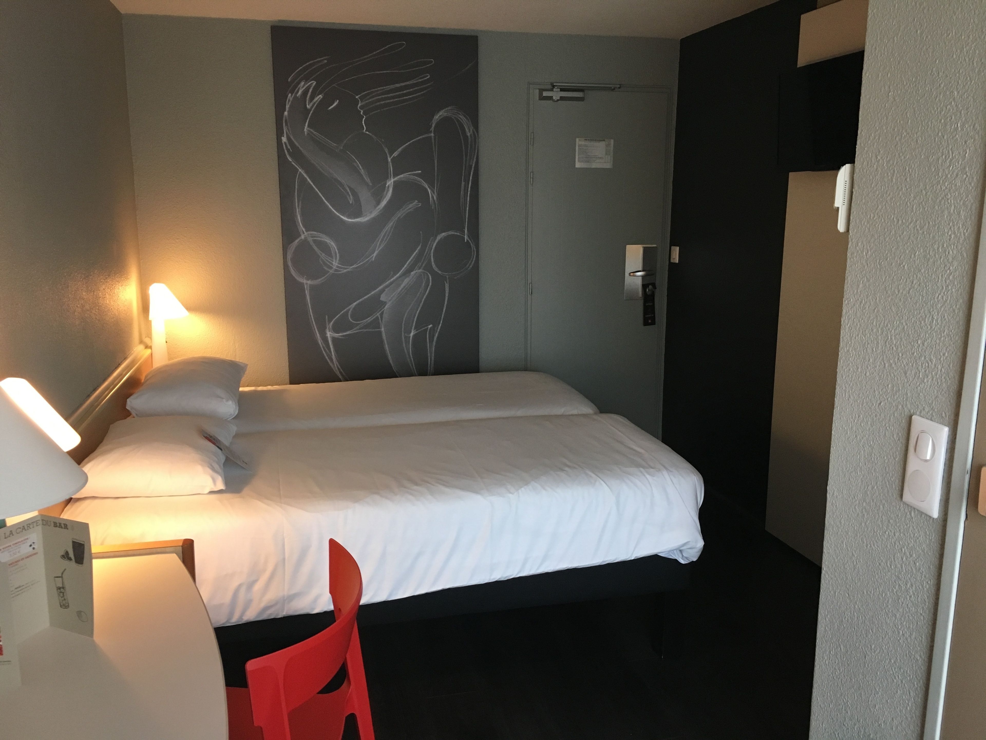 A hotel room with a bed, a desk, a chair, a wall art piece, and a door.