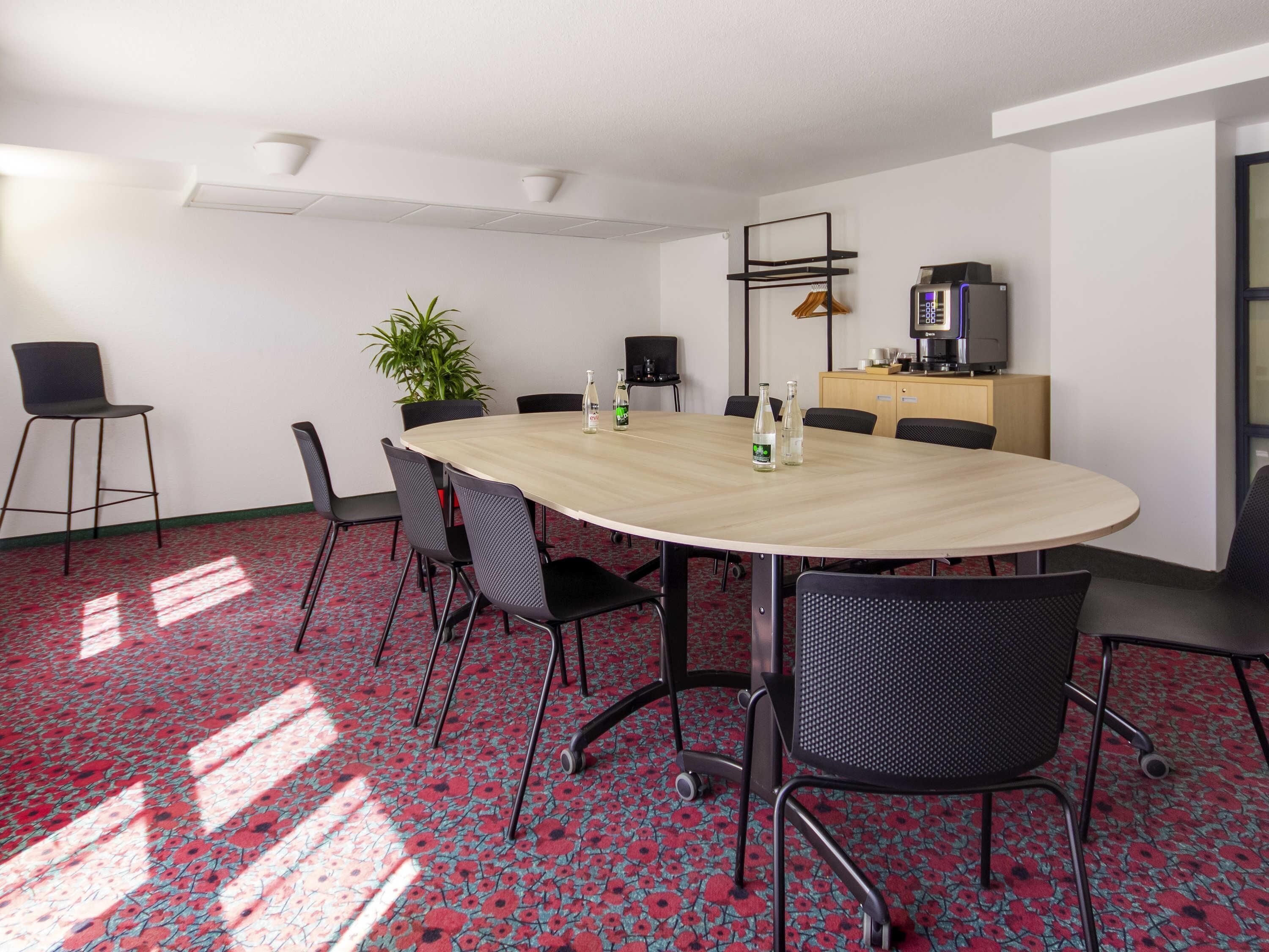 Een vergaderruimte met een ronde tafel, zwarte stoelen, een potplant, een koffieapparaat en een televisie op een kast.