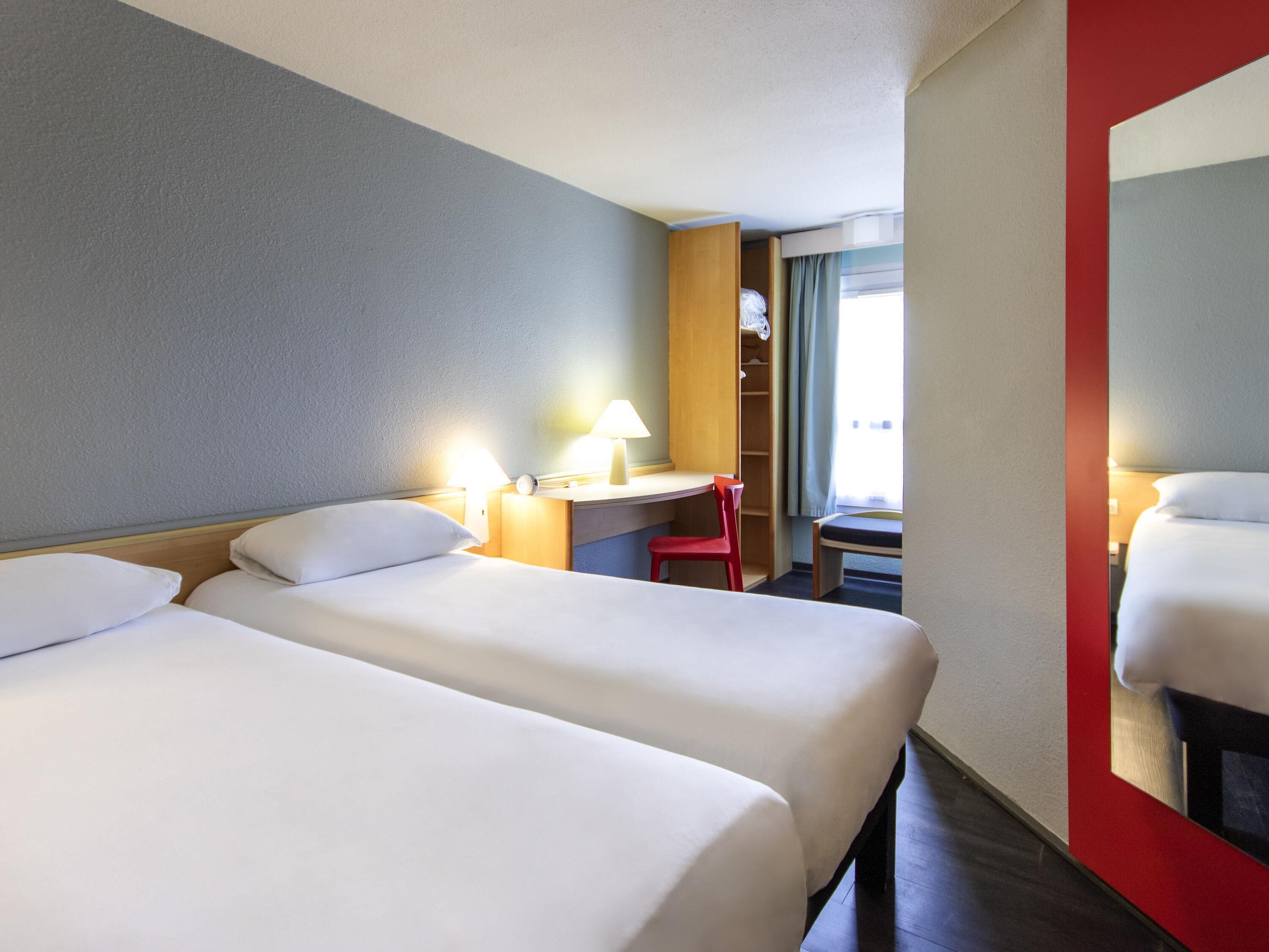Une chambre d’hôtel avec deux lits, un bureau avec une chaise, une lampe et un grand miroir.