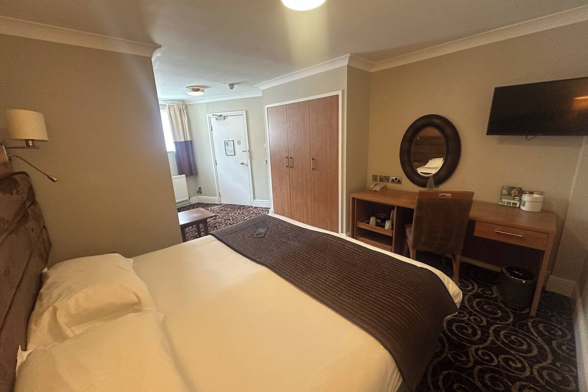 A hotel room with a large bed, a desk with a chair, a mirror, and a television on the wall.