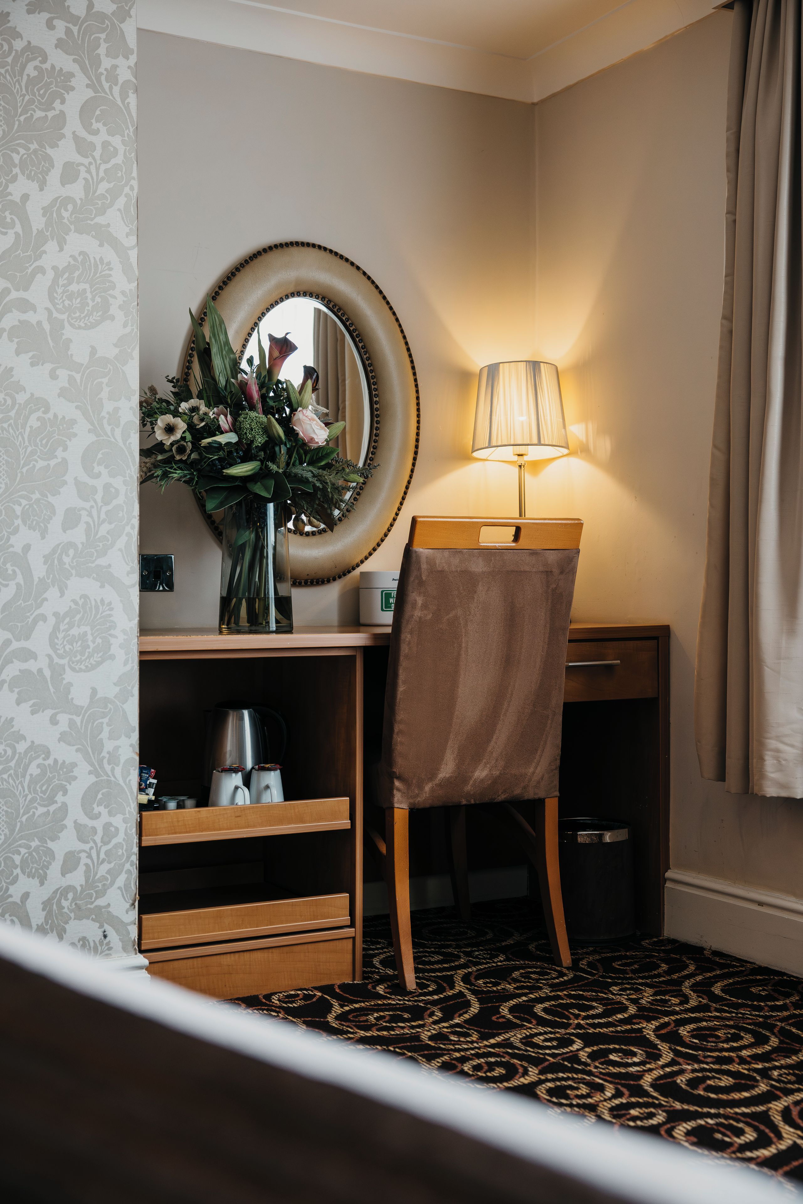 Een hotelkamer met een bureau, een lamp, een spiegel, een vaas met bloemen, een koffiezetapparaat en een prullenbak.