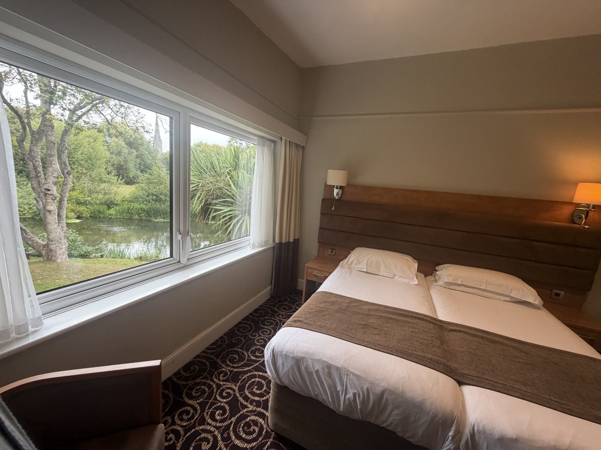 Een hotelkamer met een bed, nachtkastje, lamp en een raam met uitzicht op groen.