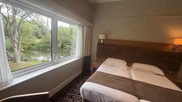 Een hotelkamer met een bed, nachtkastje, lamp en een raam met uitzicht op groen.