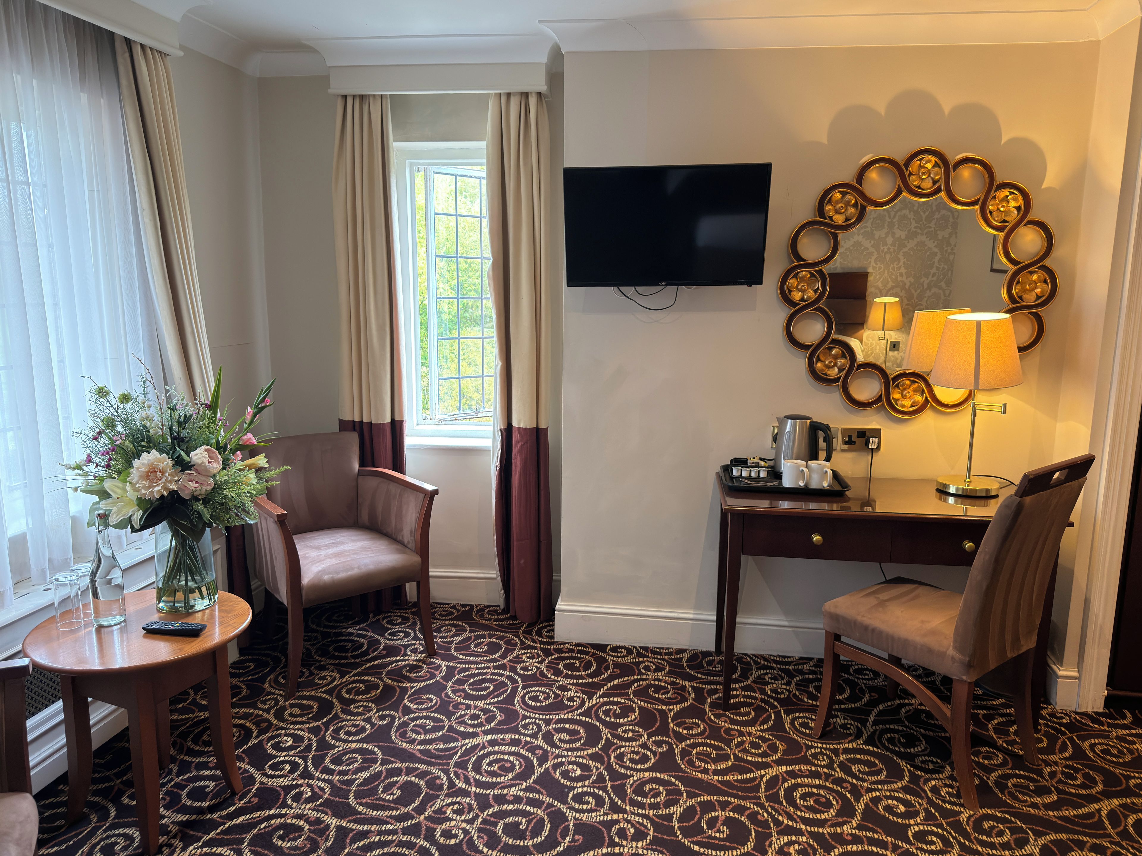 A hotel room with a desk, two chairs, a TV, a decorative mirror, a lamp, a coffee maker, and a floral arrangement.