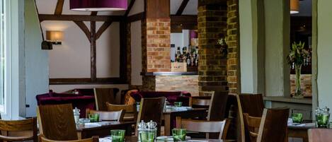 A restaurant interior with wooden tables and chairs, brick walls, and a bar area.