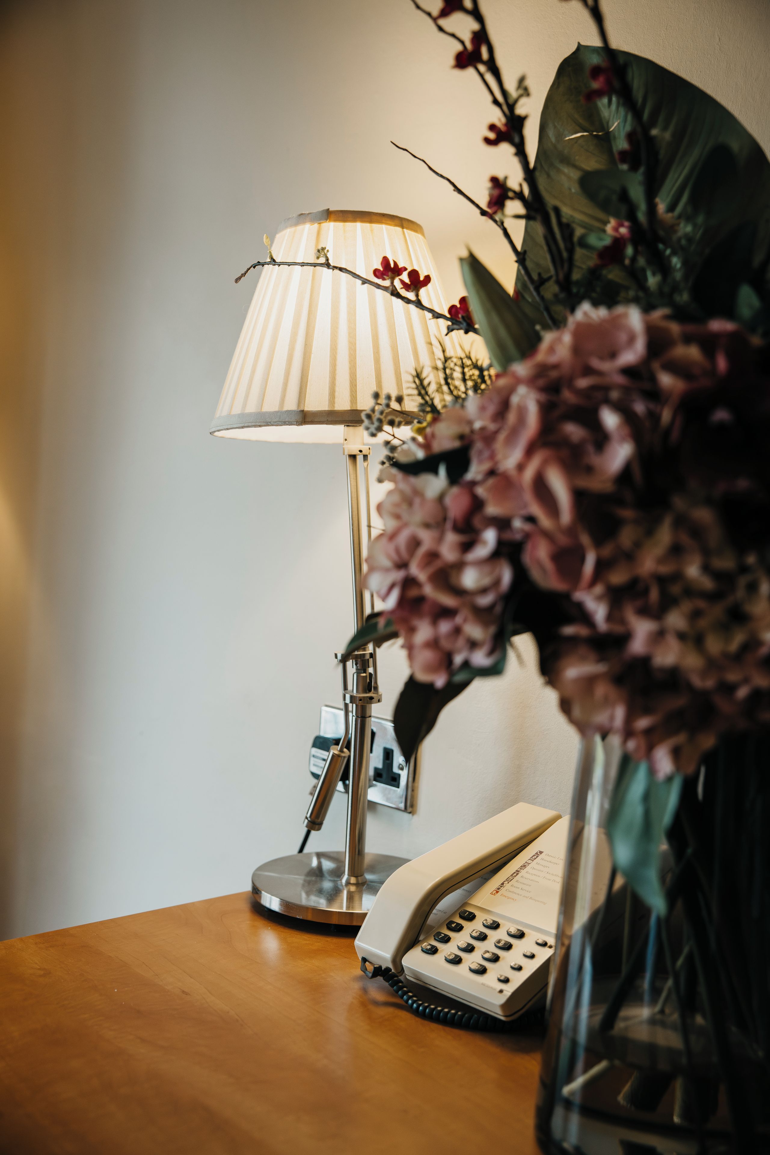 Una mesita de noche con una lámpara, un teléfono y un florero con flores.