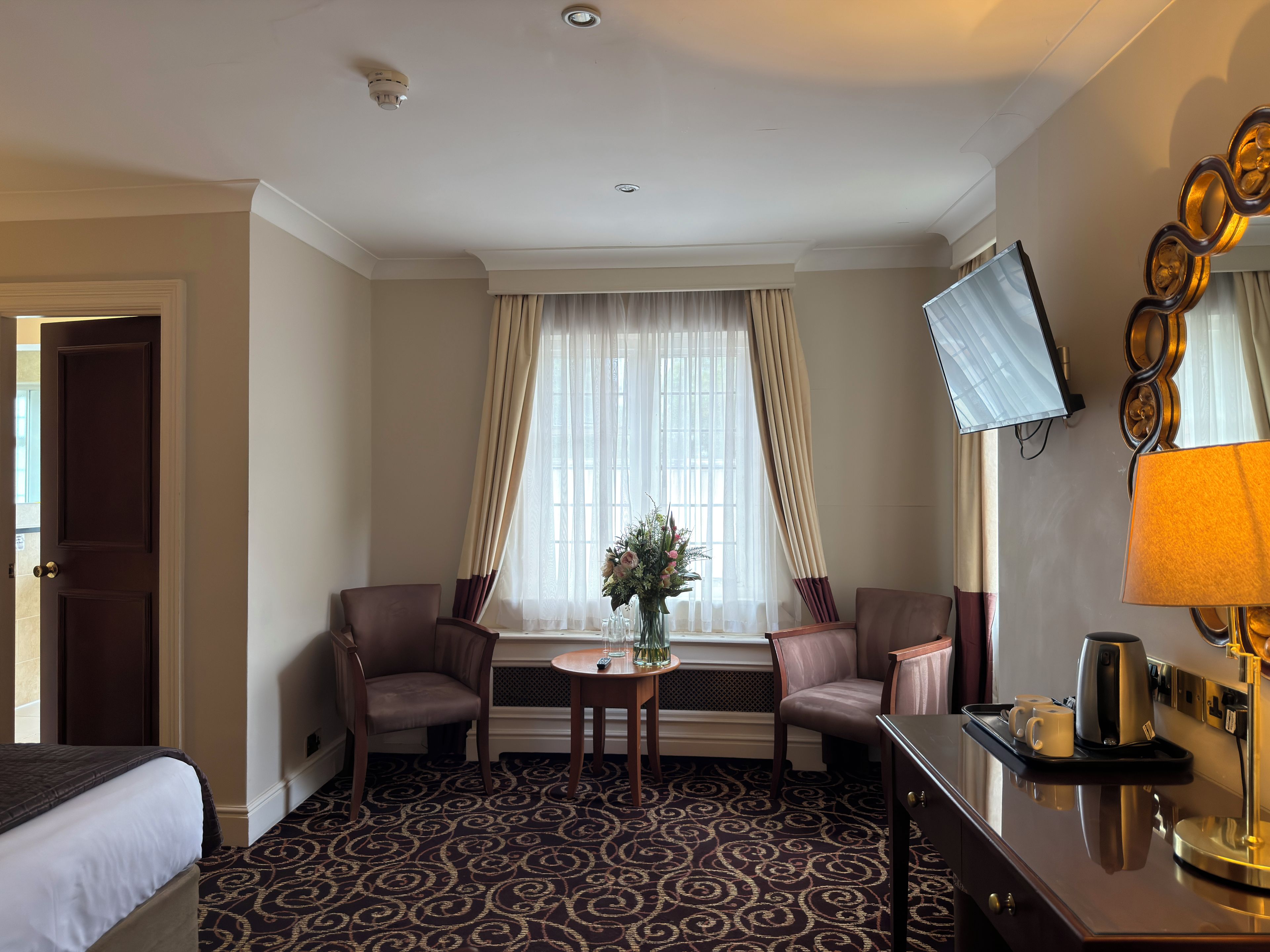 A hotel room with a bed, two armchairs, a small table with a vase, and a desk with a lamp and coffee maker.