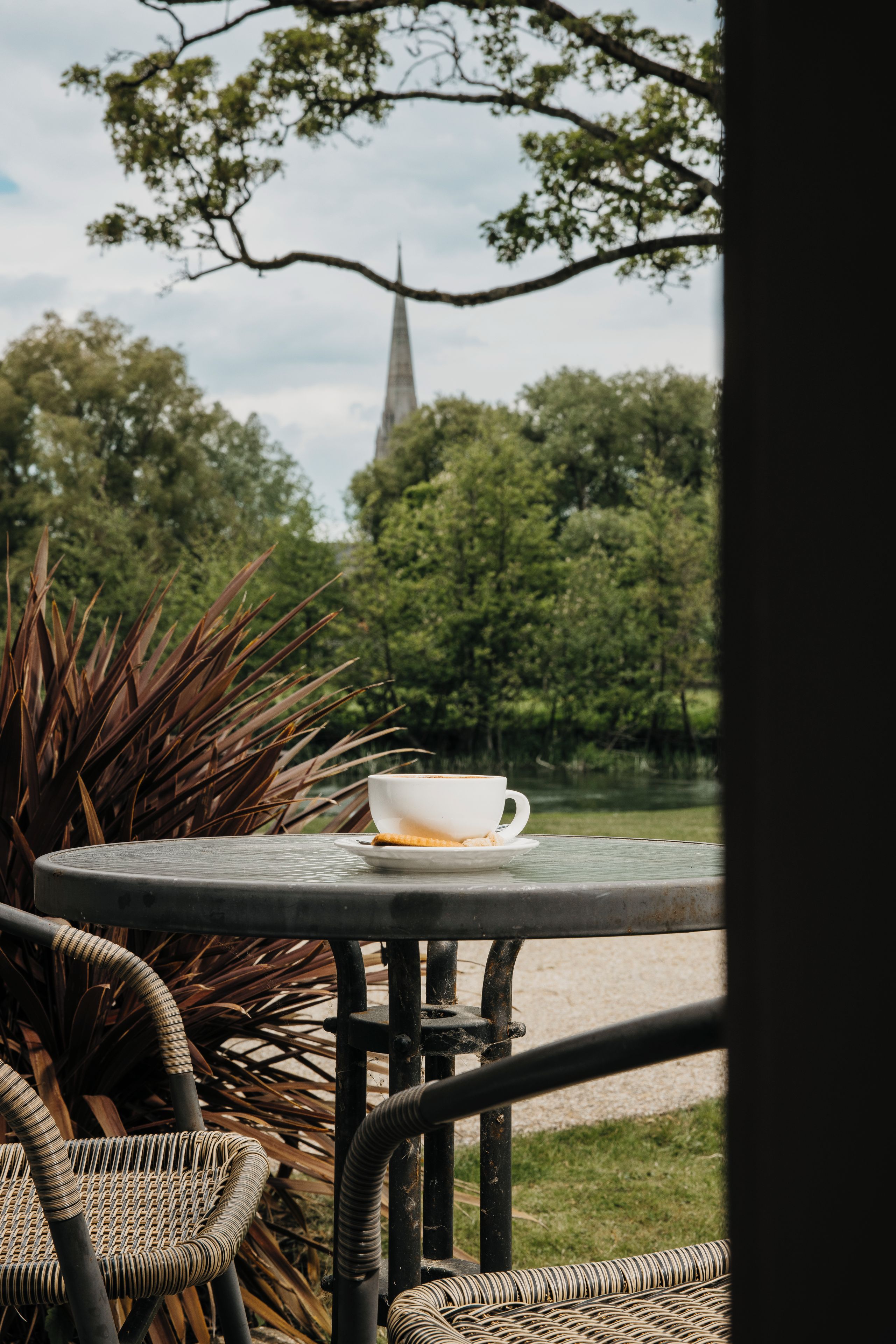 A table with a cup and saucer, wicker chairs, and a view of a church spire through trees.