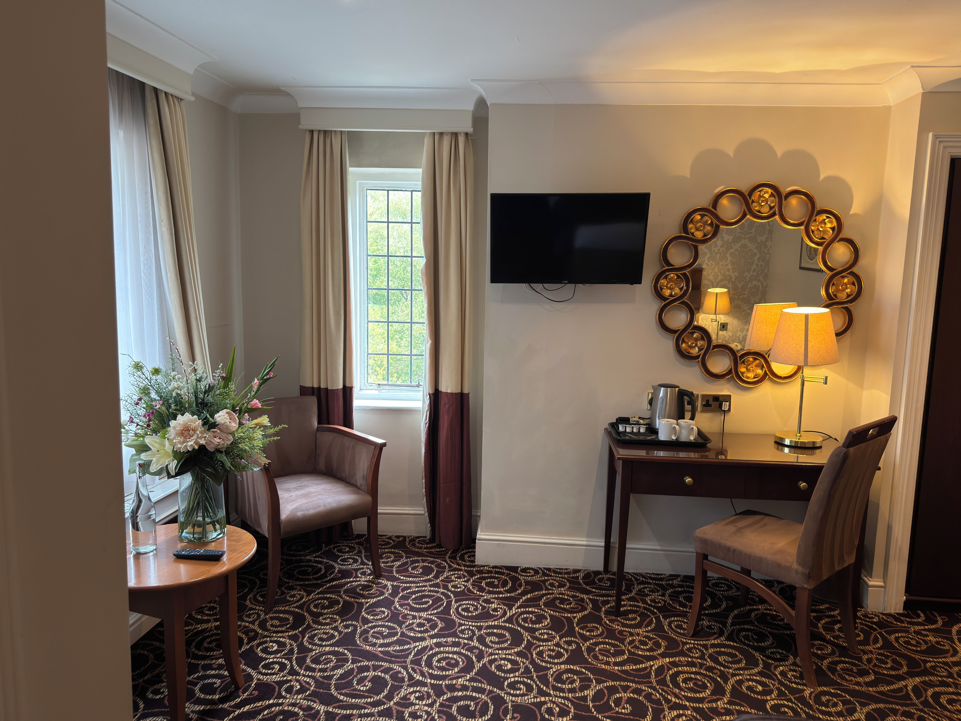 Une chambre d’hôtel équipée d’une télévision, d’un bureau avec une lampe, d’une chaise, d’une petite table ornée d’un vase de fleurs et d’un tapis à motifs.