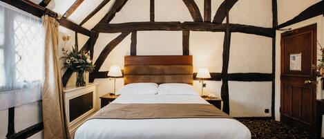 A traditional room with exposed wooden beams, a large bed with white linens, a nightstand with a lamp, and a window with curtains.