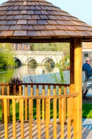 A gazebo with a tiled roof overlooking a river, a stone bridge, and a building in the background.