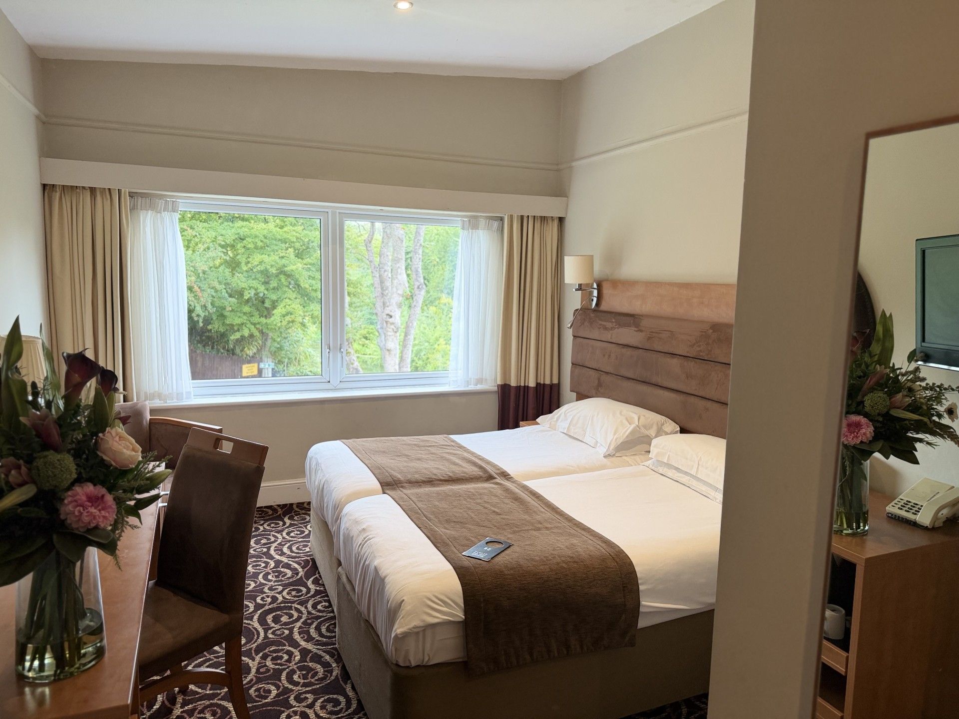 A hotel room with a large bed, a desk with a chair, a window with curtains, and a vase with flowers.