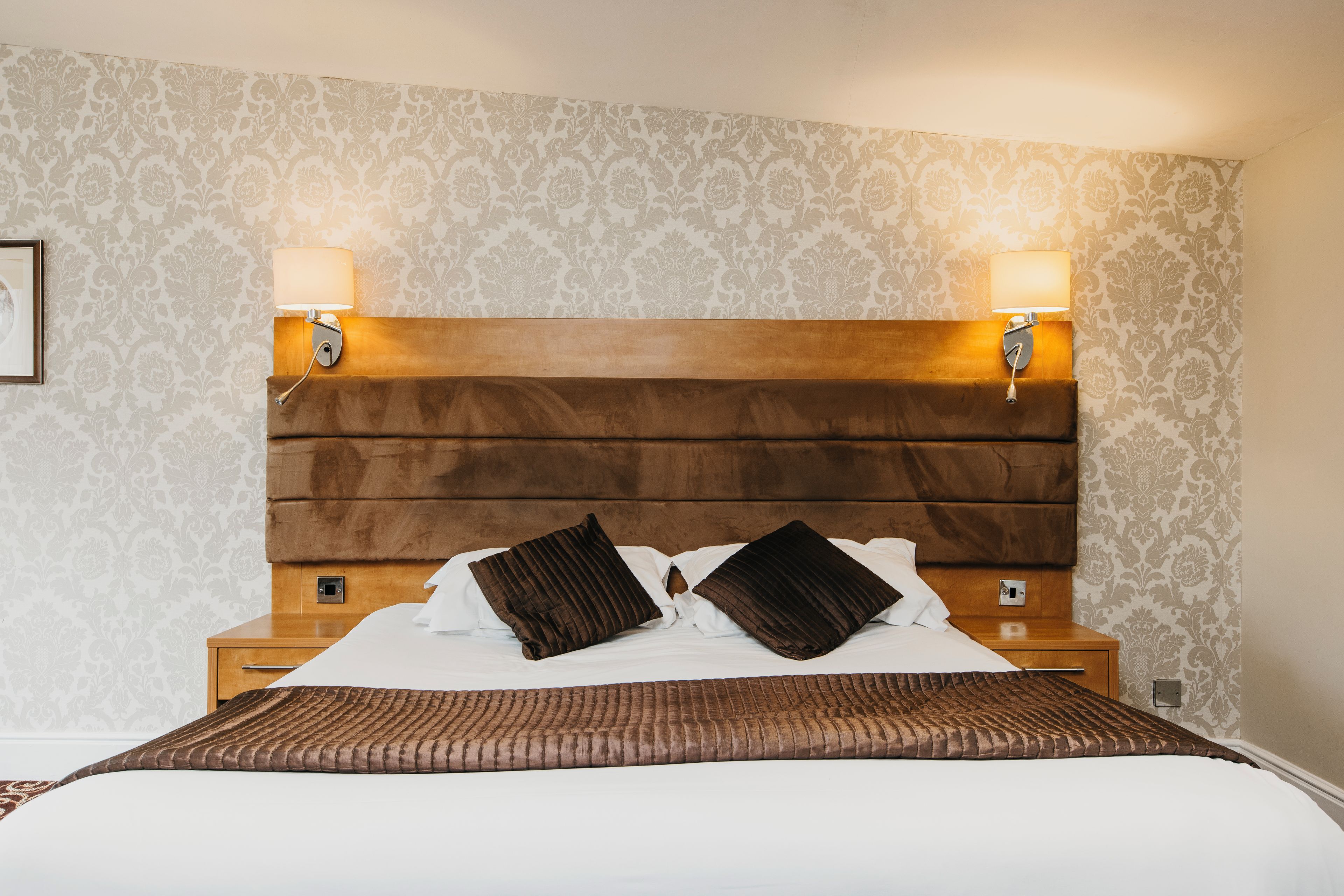 A hotel room with a large bed, two bedside lamps, a wooden headboard, and patterned wallpaper.
