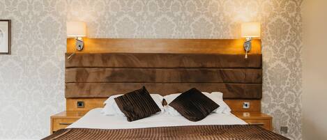 A hotel room with a large bed, two bedside lamps, a wooden headboard, and patterned wallpaper.