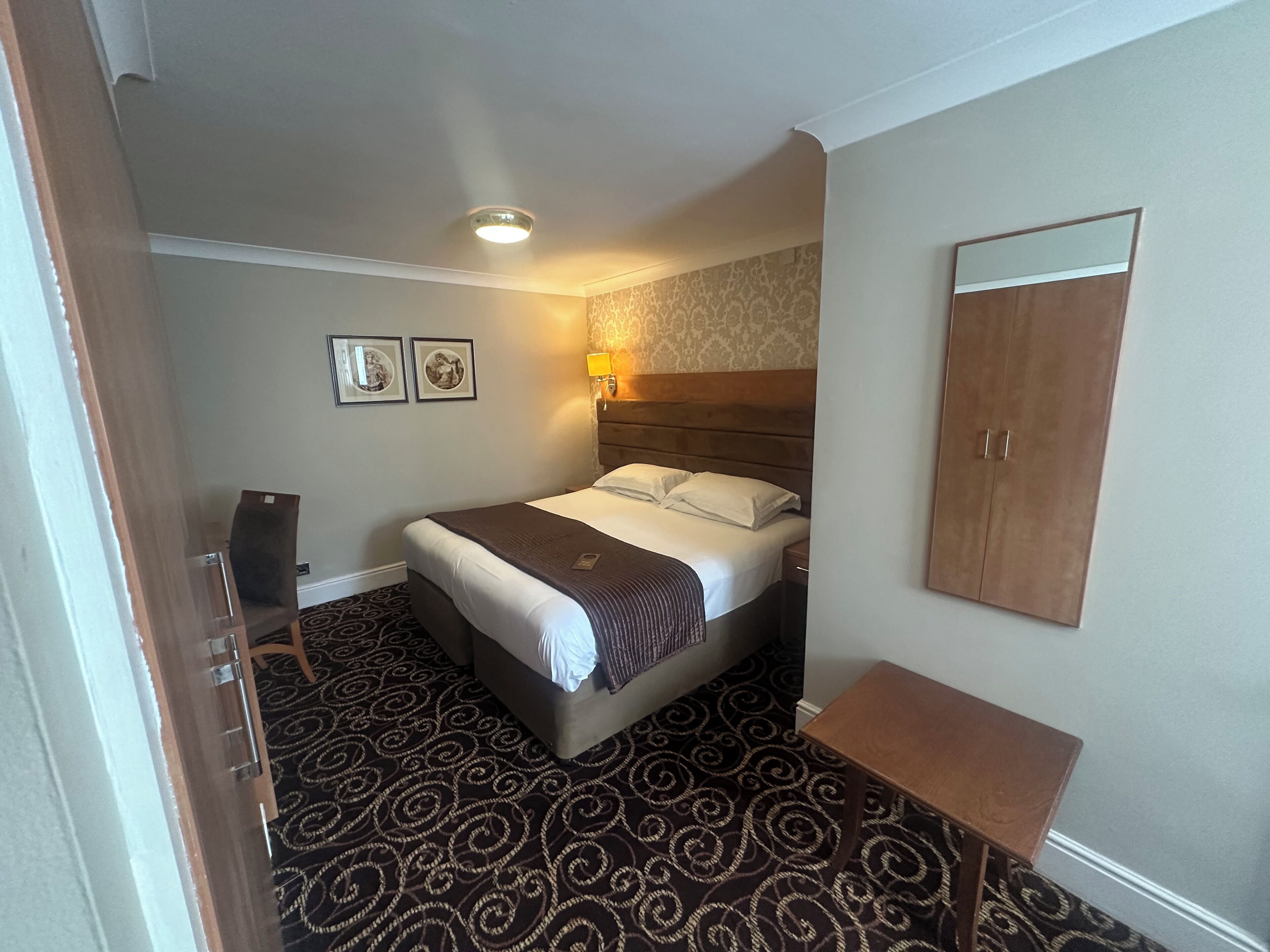 A hotel room with a bed, a nightstand, a desk, a wardrobe, and a patterned carpet.