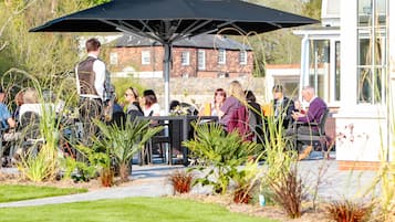 Een terras met tafels en stoelen onder een grote parasol, omringd door planten en een bakstenen gebouw op de achtergrond.