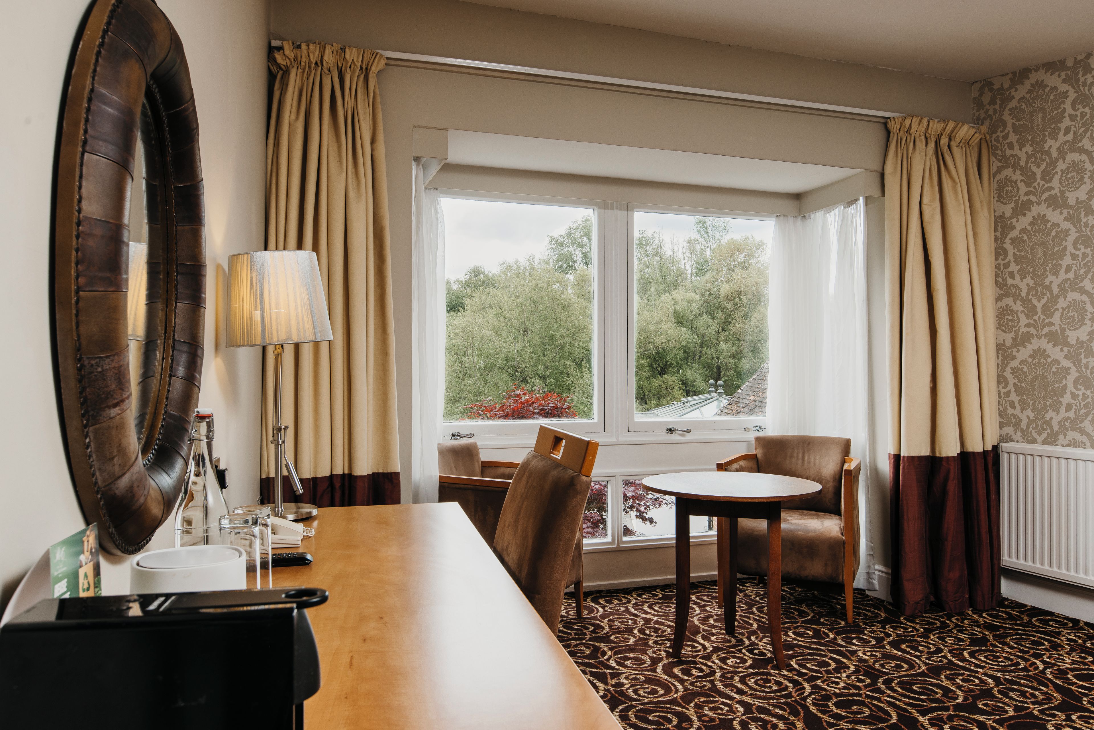 A hotel room with a dining table, chairs, a lamp, a mirror, and a view of greenery outside.