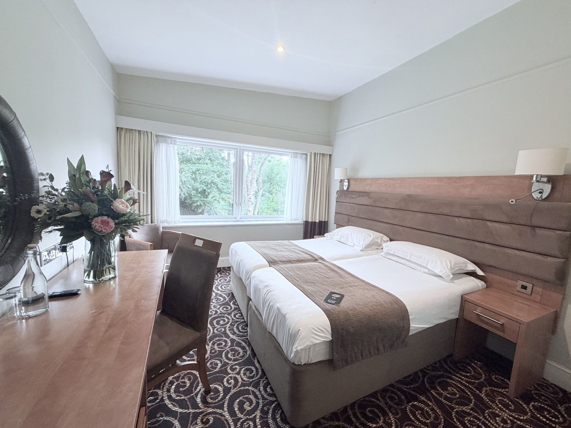 Una habitación de hotel con cama, mesita de noche, lámpara y ventana con cortinas.