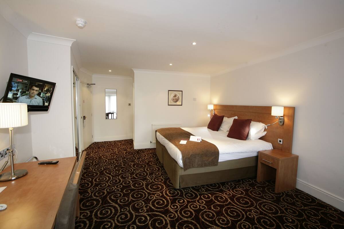 A hotel room with a bed, bedside table, lamp, and a television displaying a live broadcast.